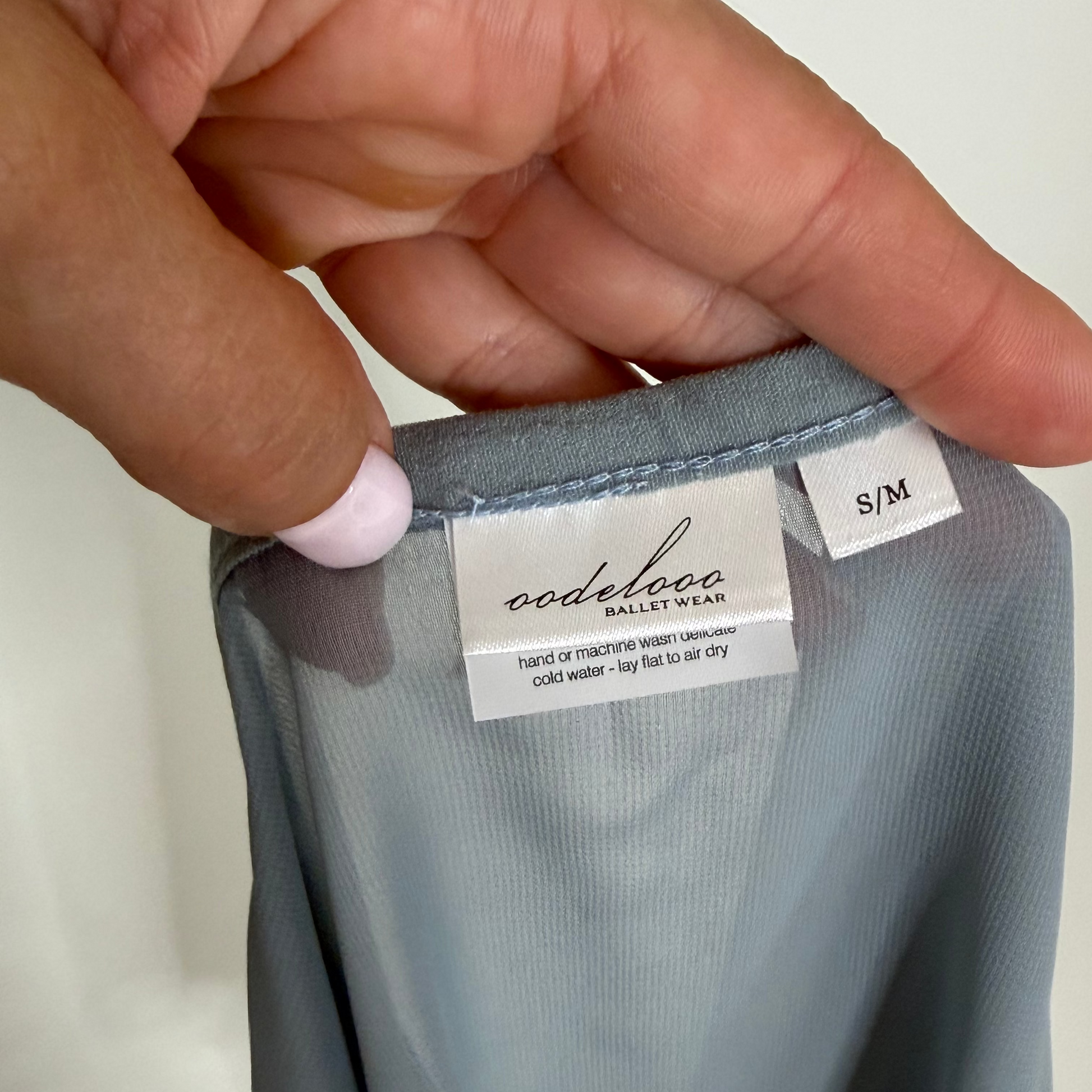 A close-up of a hand holding the waistband of a slate blue-gray dress, showing the 'oodelooo ballet wear' brand tag and an 'S/M' size label.