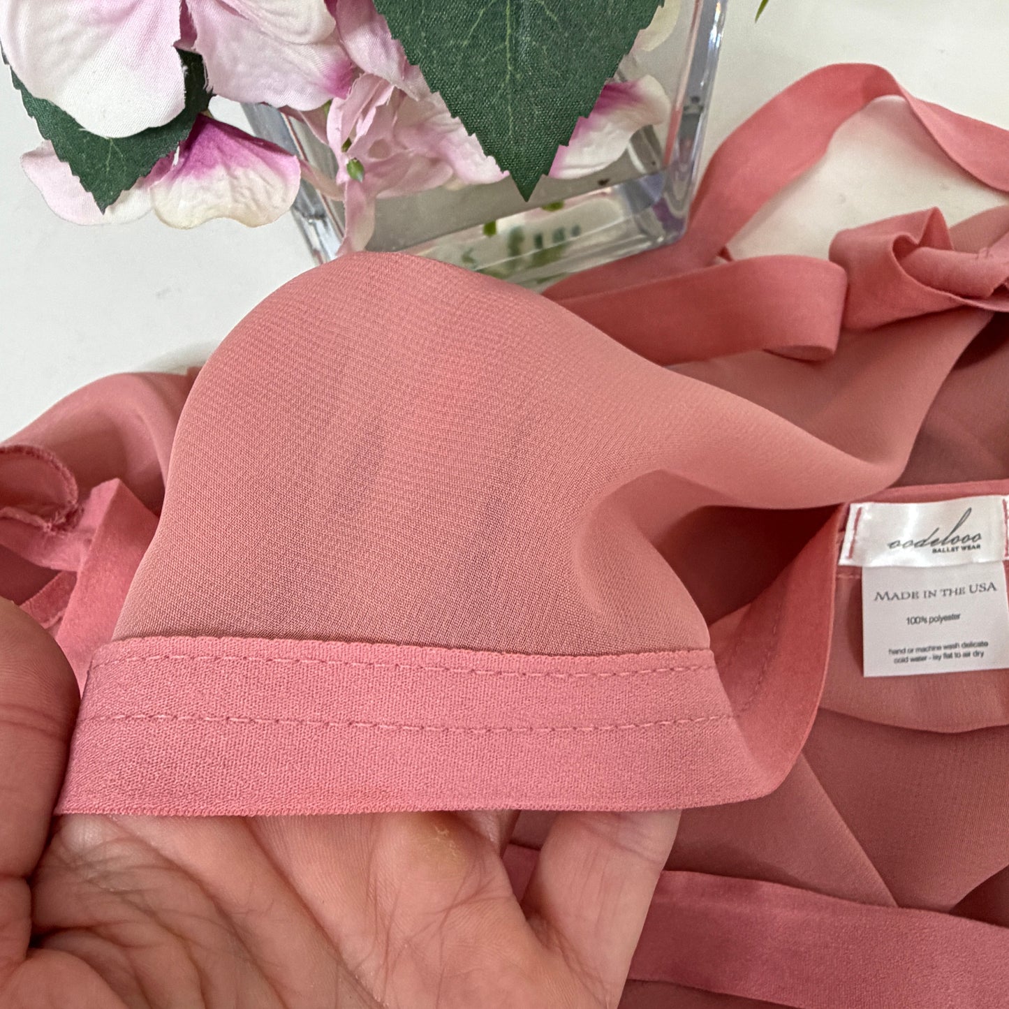 Close-up of pink fabric with a visible brand label held by a hand, with flowers in the background.