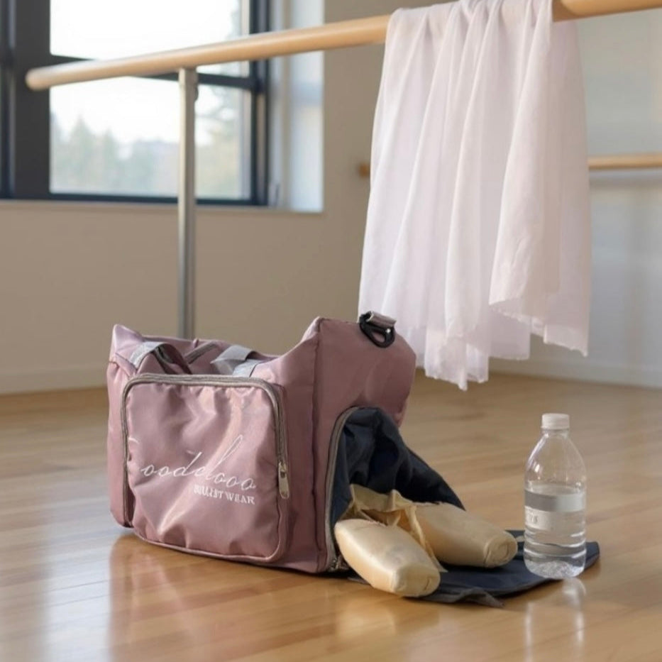 Pink dance bag with ballet shoes and water bottle on a wooden floor in a studio.